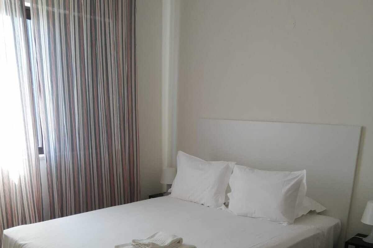 Una cama bien hecha con sábanas y almohadas blancas, una toalla doblada sobre la cama, suelo de madera y una cortina a rayas parcialmente abierta.