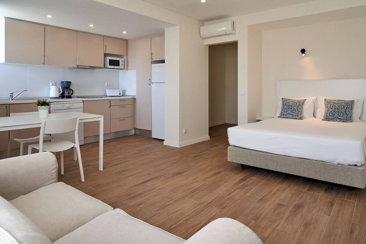 Une chambre d'hôtel compacte comprenant un coin cuisine, un lit simple avec des coussins à motifs et un petit coin salon avec un canapé et des fauteuils.