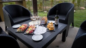 A dining table set outdoors with wicker chairs, plates, glasses, and a bottle of wine.