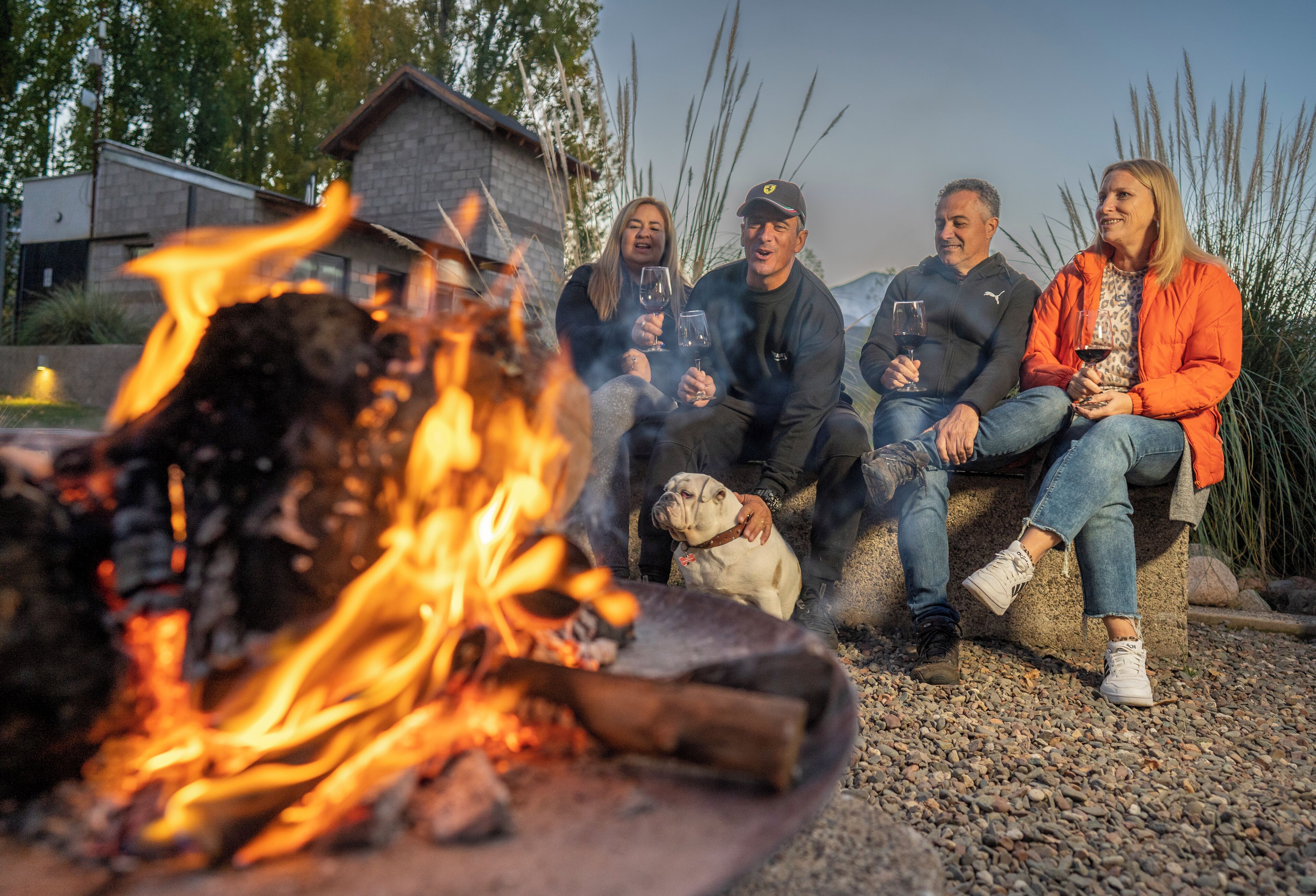 A group of people sitting around a campfire with a dog, in a rustic outdoor setting.