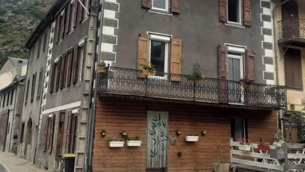 Un bâtiment à deux étages doté d’une façade en bois, de nombreuses fenêtres avec volets et d’un balcon agrémenté de plantes en pot.