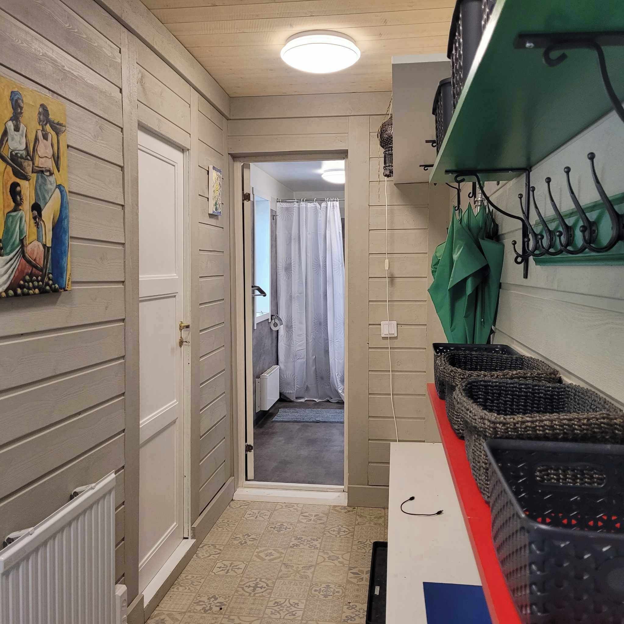 A narrow hallway with a door leading to a bathroom. The hallway has a radiator, a painting on the wall, and a green umbrella rack with hooks.