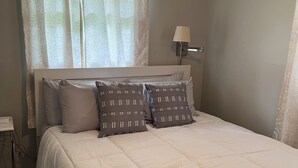 Uma cama bem arrumada com edredom branco e dois travesseiros, abajur e janela com cortinas leves.