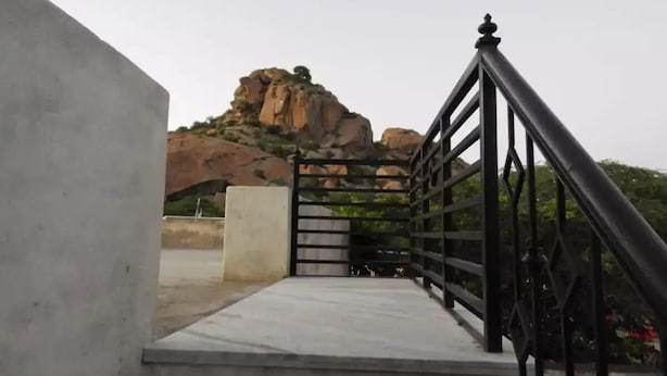 Una escalera de concreto que conduce a una reja con baranda de metal, rodeada de terreno rocoso y vegetación escasa.