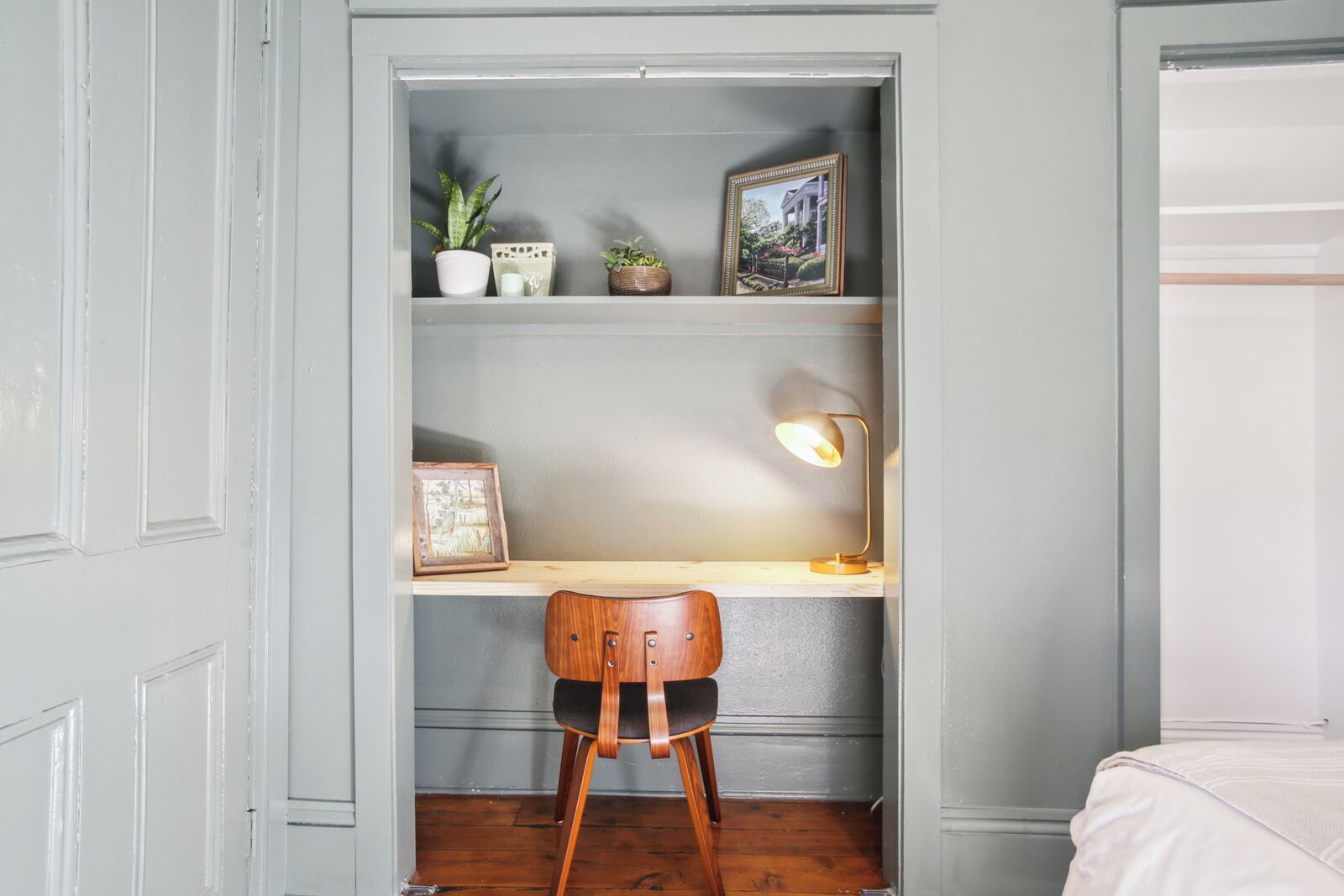 A study with a wooden chair, a desk, a lamp, and shelves with plants and framed pictures.