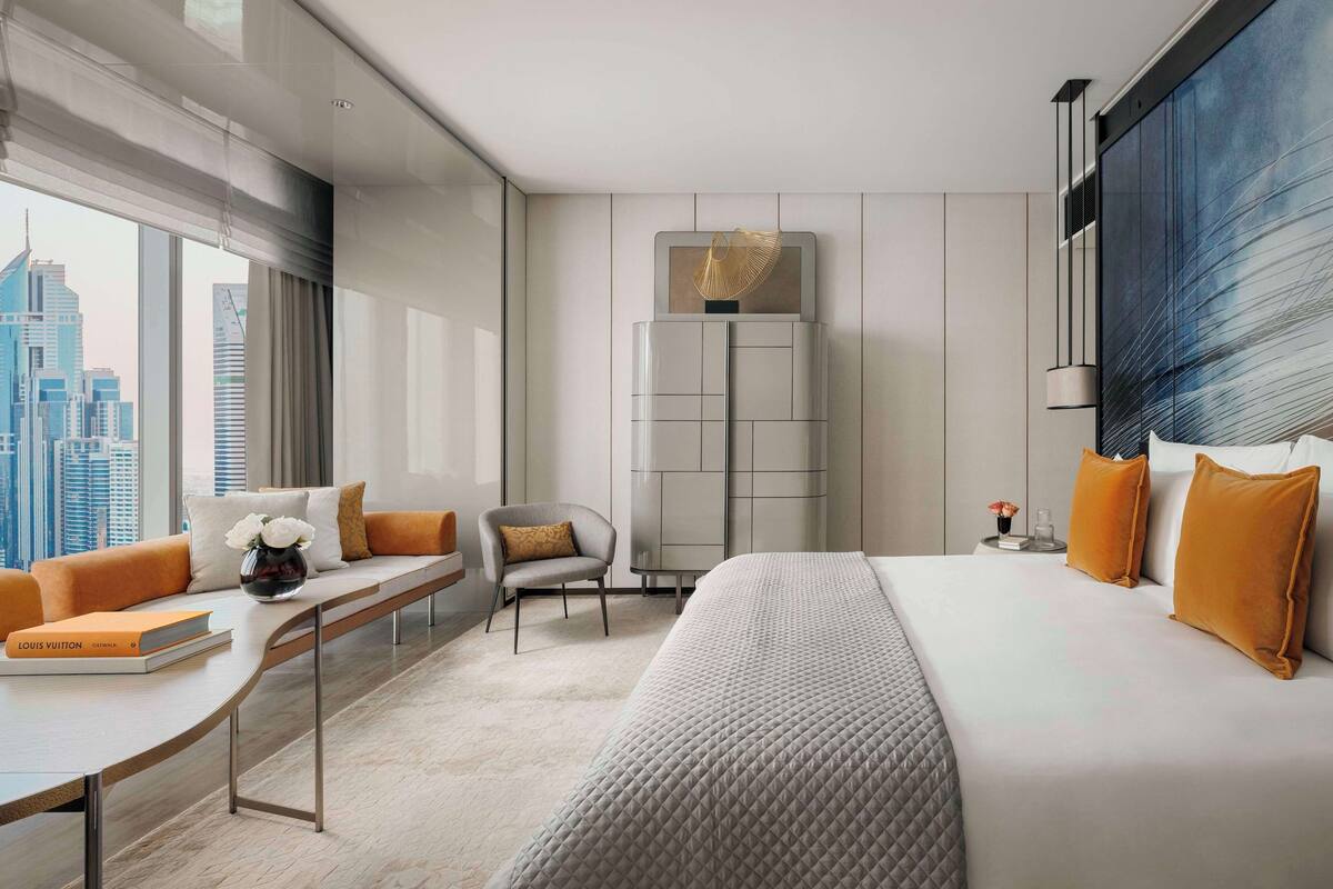 Une chambre d’hôtel moderne dotée d’un grand lit, d’un coin salon avec un canapé et un fauteuil, d’une petite table et offrant une vue sur le panorama urbain grâce à de grandes fenêtres.