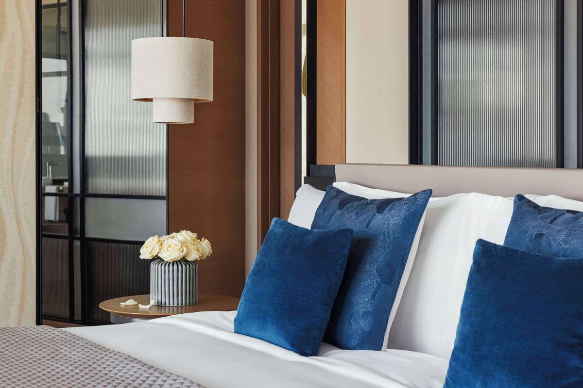 Un lit bien fait, avec des oreillers bleus et blancs, un vase rempli de fleurs blanches et une lampe fixée au mur.