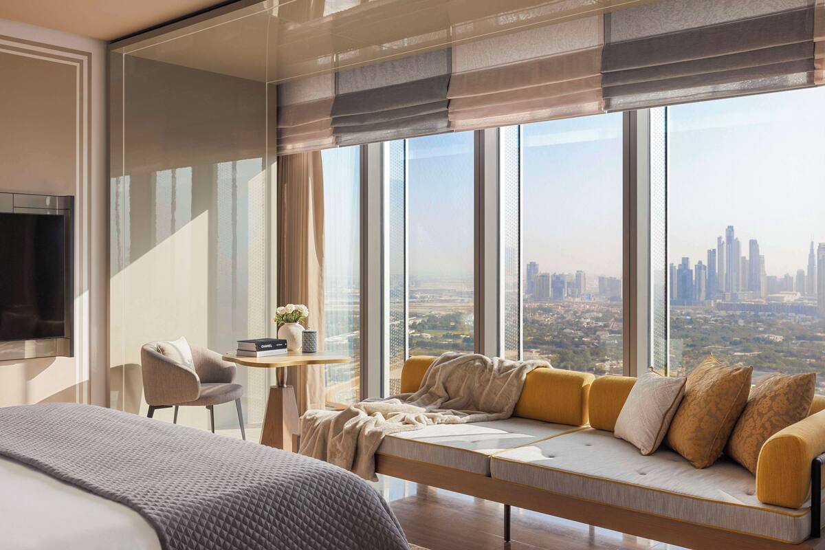 Une chambre d’hôtel moderne avec un grand lit, un canapé avec des coussins, une petite table avec un vase et une vue sur le paysage urbain à travers de grandes fenêtres.