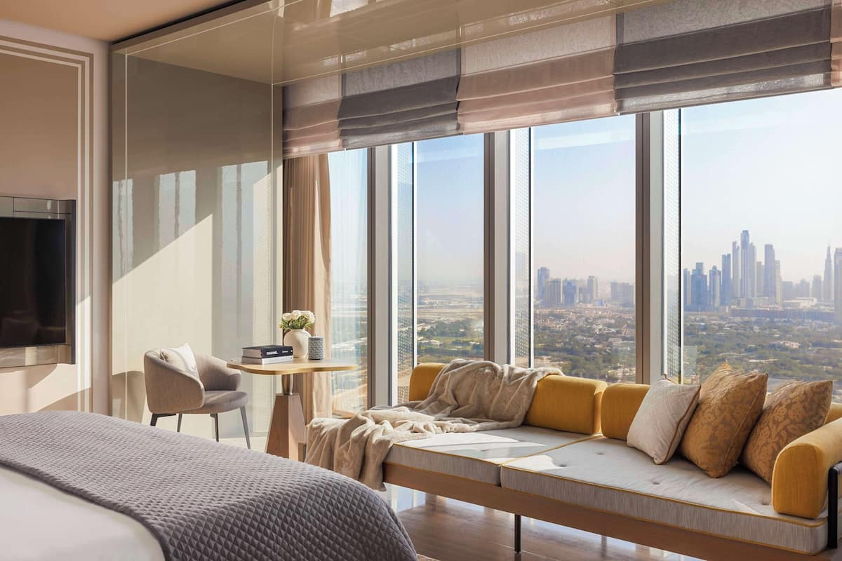 A modern hotel room with a large bed, a sofa with cushions, a small table with a vase, and a view of a cityscape through large windows.