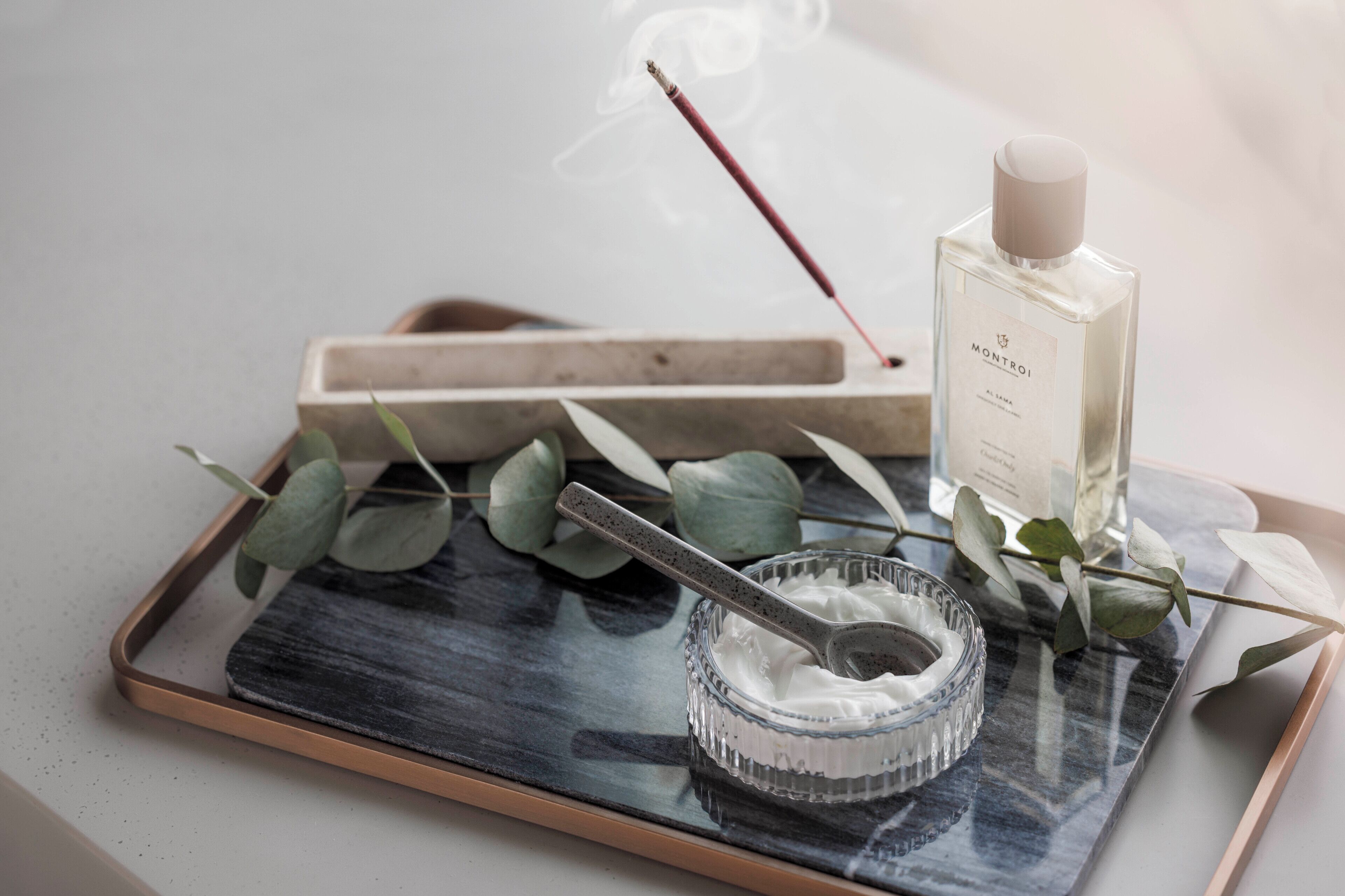 A tray with a bottle of Montroi cologne, a small bowl of cream, eucalyptus leaves, and a smoking incense stick.