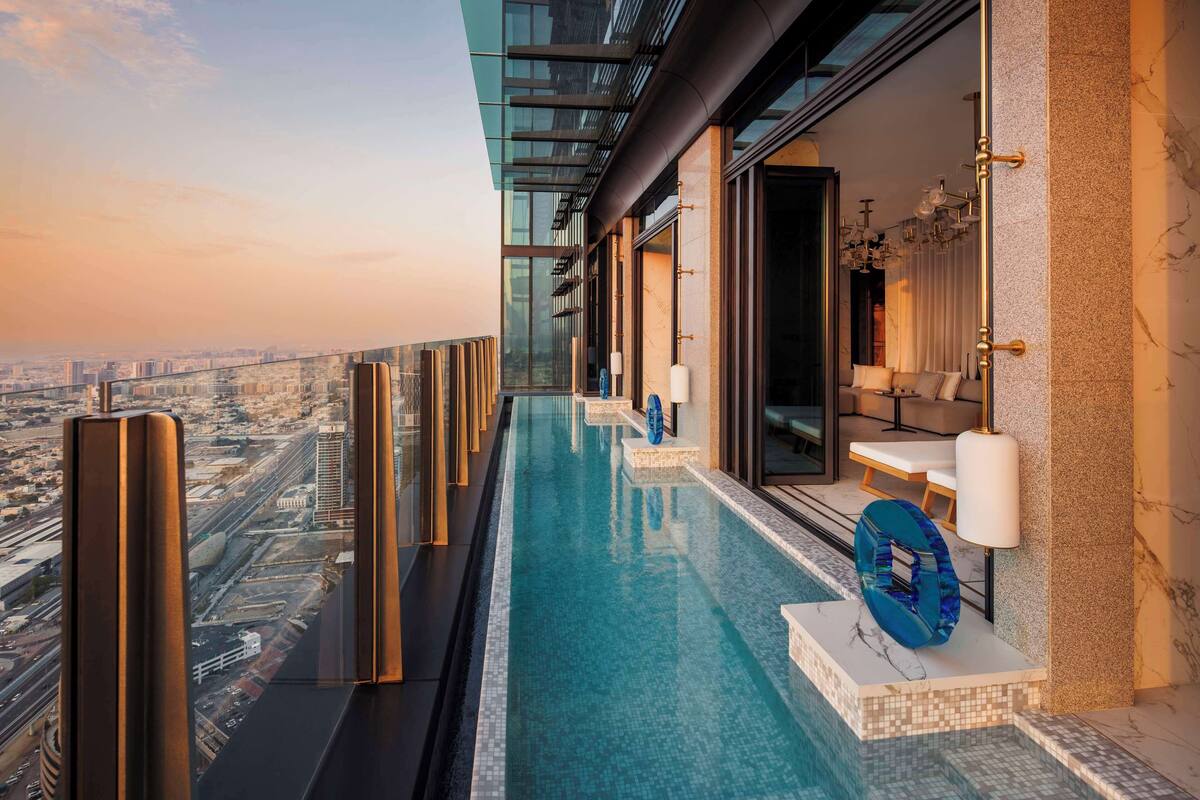 Une piscine à débordement située sur le toit, offrant une vue sur la ville, avec une balustrade en verre et un espace détente comprenant un canapé et des lampes.