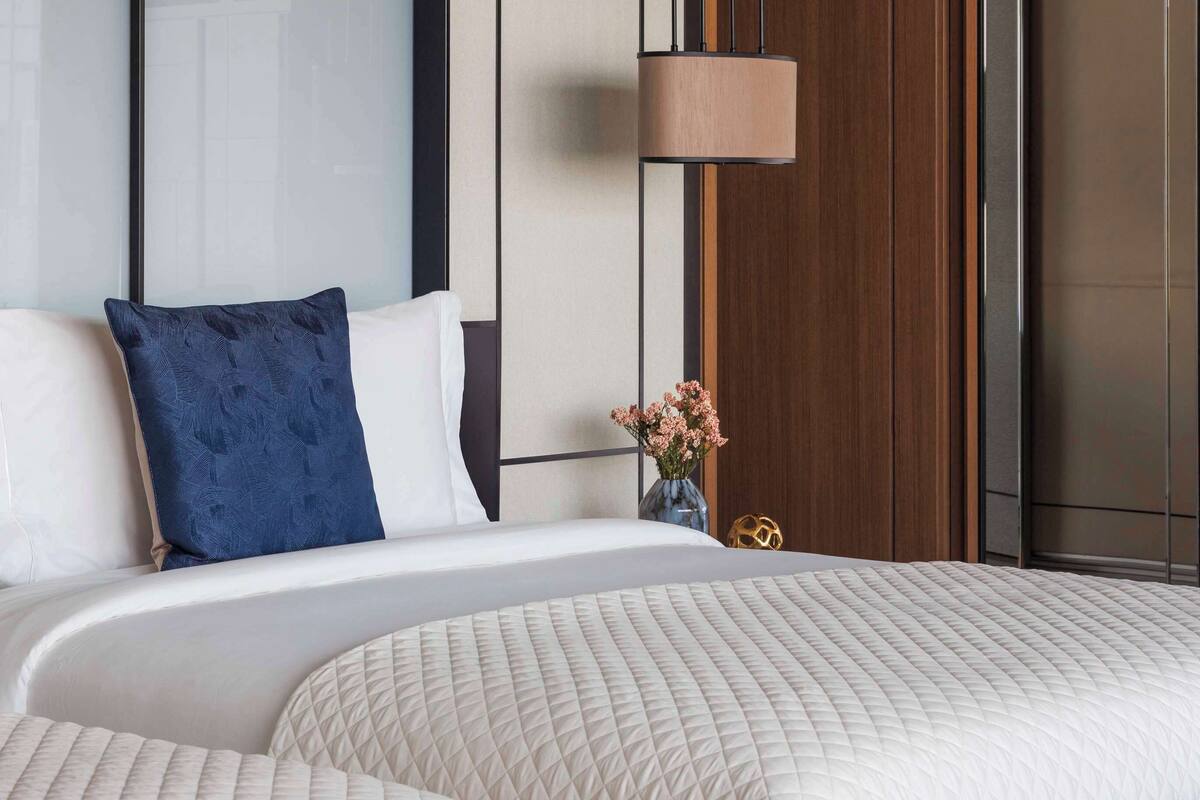 Une chambre d’hôtel moderne avec un grand lit, une table de chevet ornée d’un vase de fleurs et une grande fenêtre donnant sur la ville.