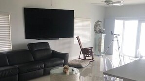 A modern living room with a black sofa, a round coffee table, and a TV mounted on the wall. There are ceiling fans, a window with blinds, and a clear view of the outside. - Breathtaking sunrises from your balcony at this 3 bedroom, 2 bath home (Big Pine Key)