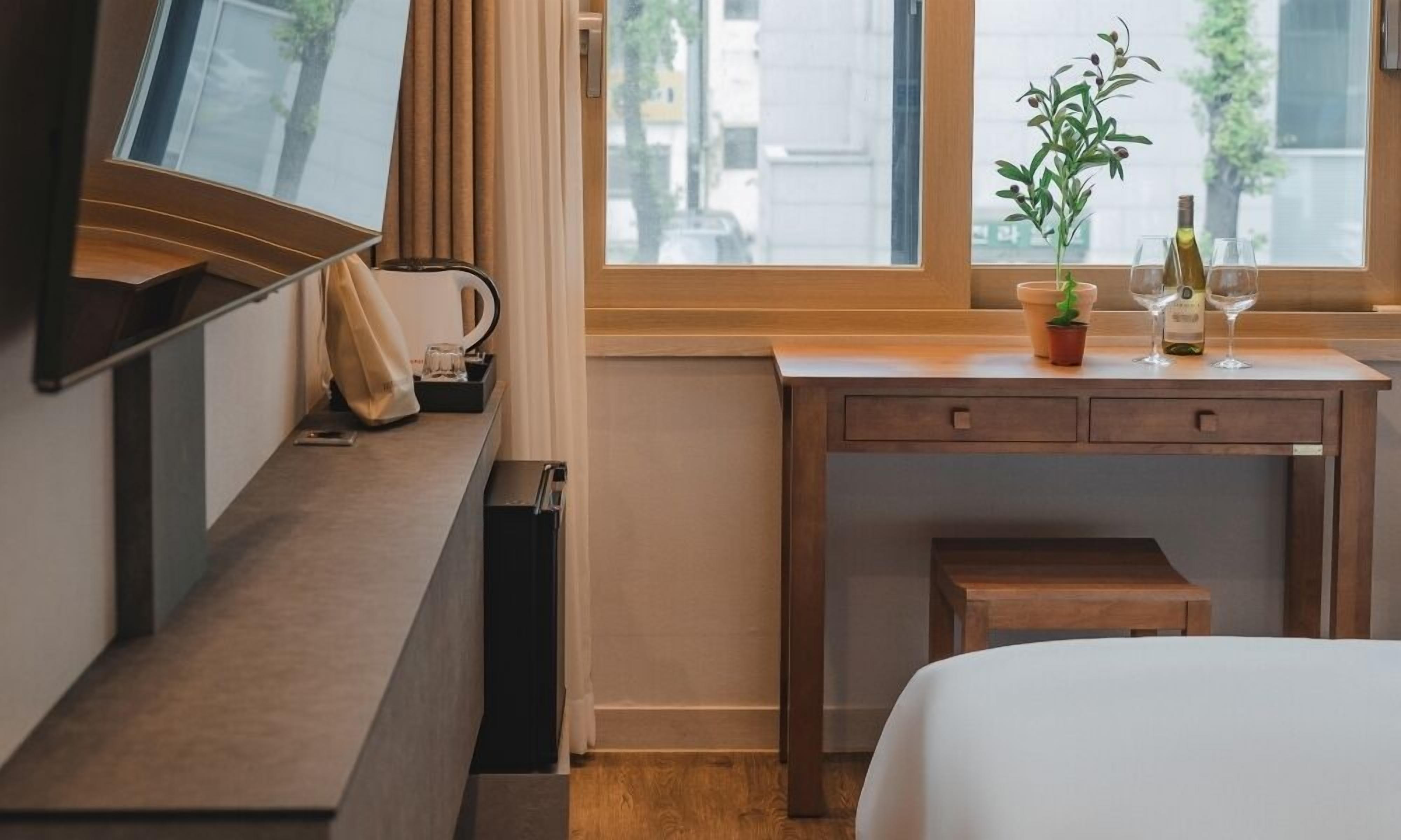 A hotel room with a wooden desk, a bed, a TV, a window with curtains, and a small potted plant.