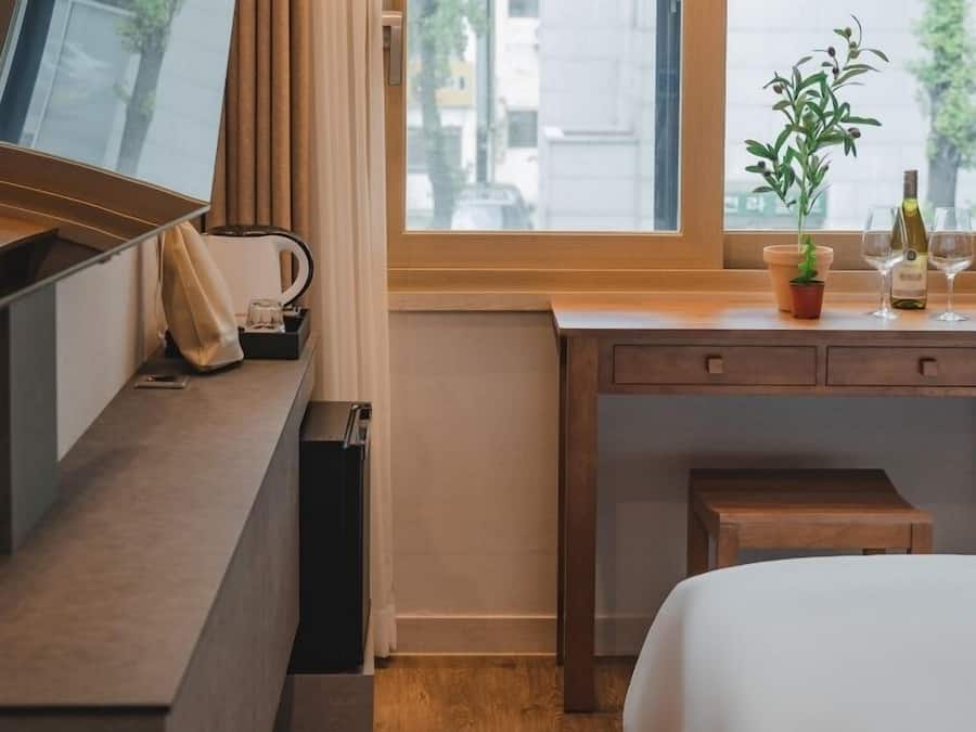 A hotel room with a wooden desk, a bed, a TV, a window with curtains, and a small potted plant.