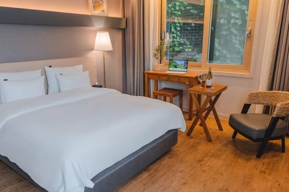 Ein Hotelzimmer mit einem großen Bett, einem Schreibtisch mit Computer, einem Sessel und einem Fenster mit Blick auf Bäume.