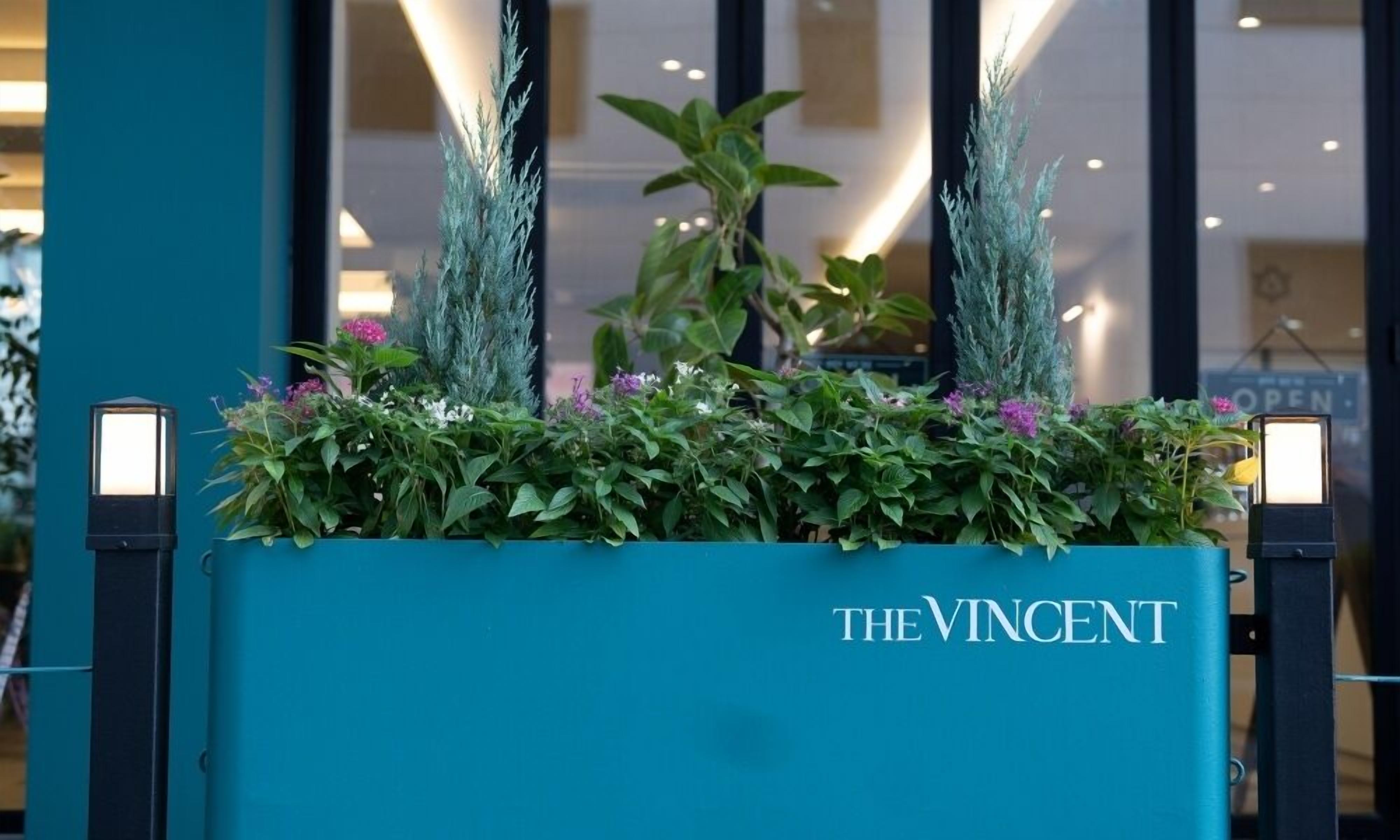 A teal planter with a sign reading "THE VINCENT" in front of a building with vertical windows.