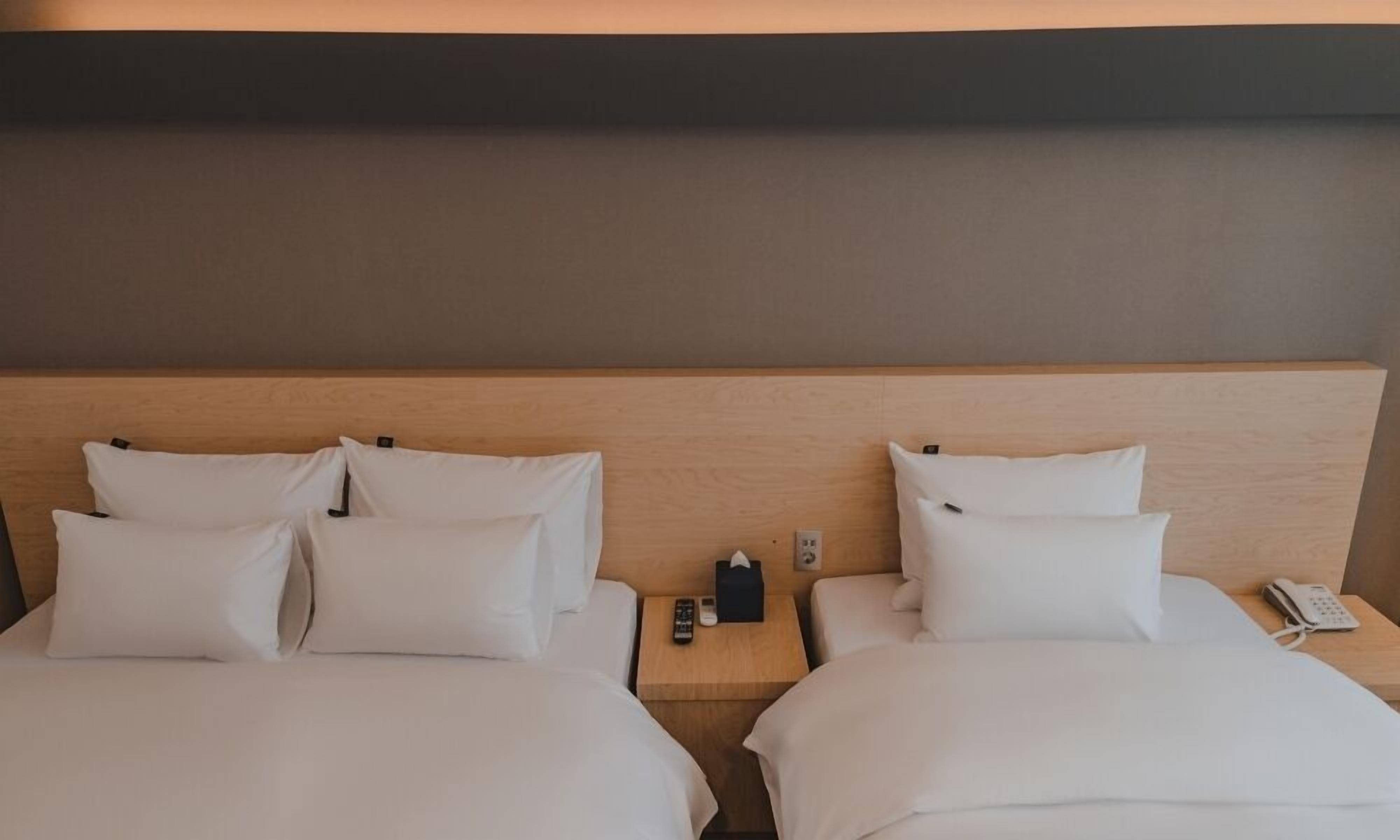 A hotel room with two beds, a wooden headboard, a bedside table with a phone and remote control, and a wall socket.