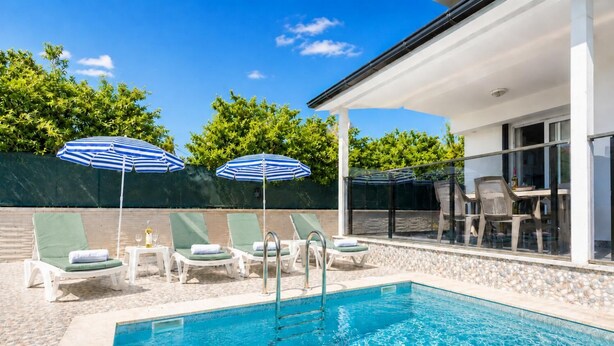 En swimmingpool med liggestole og parasoller, et overdækket siddeområde og udsigt til en bygning med store vinduer.