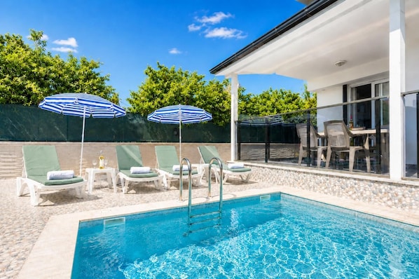 Une piscine dotée de chaises longues et de parasols, un espace de détente couvert et une vue sur un bâtiment aux grandes fenêtres.