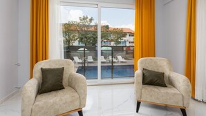 Une pièce comprenant deux fauteuils, une grande porte-fenêtre coulissante donnant sur une zone abritant une piscine, et offrant une vue sur les bâtiments et les arbres à l’extérieur.