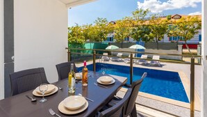 Un coin repas avec une table dressée pour deux personnes, donnant sur une piscine et offrant une vue sur des bâtiments et des arbres.