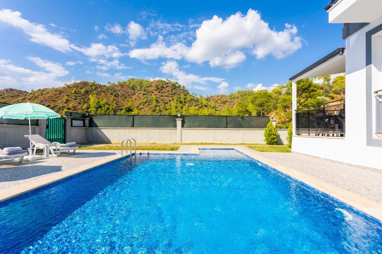 A swimming pool with lounge chairs and an umbrella, surrounded by a white building and a green fence.