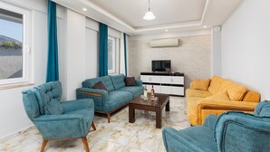 Una sala de estar con sofás verde azulado y mostaza, una mesa de centro y un televisor sobre un mueble blanco.