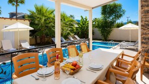 Área de refeições junto à piscina, com uma mesa posta para dois, rodeada por espreguiçadeiras e guarda-sóis, com vista para a piscina.