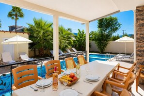 Zona de comedor junto a la piscina con una mesa para dos, rodeada de sillones de descanso y sombrillas, con vista a una alberca.