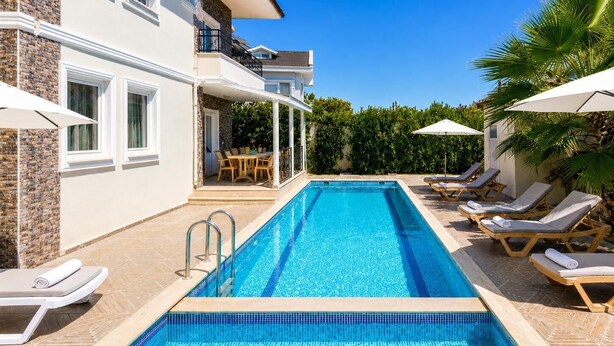Piscina com espreguiçadeiras e guarda-sóis, alpendre coberto com mesa e cadeiras, e edifício com fachada de pedra.