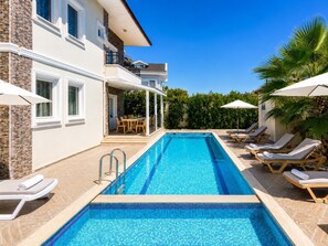 A swimming pool with lounge chairs and umbrellas, a covered patio with a table and chairs, and a stone facade building.
