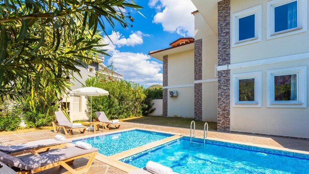 A swimming pool with lounge chairs and umbrellas, surrounded by greenery and a building with multiple windows. - Villa Kaya 2 - Five Bedroom Villa, Sleeps 12 (Dalyan)