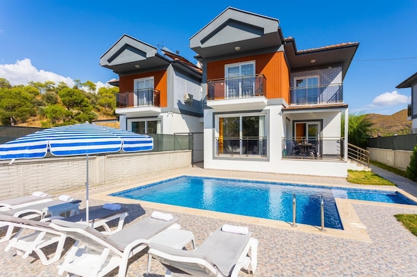 Ein modernes, zweistöckiges Haus mit Swimmingpool, Liegestühlen und einem blau gestreiften Sonnenschirm.