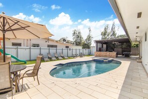 A backyard with a swimming pool, lounge chairs, a table, and a barbecue area.