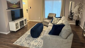 A living room with a flat-screen TV, a sofa, a chair, a ceiling fan, and a large window with curtains. - Round Rock Supernova minutes from Kalahari, Round Rock Sports Complex, and DT  (Round Rock)
