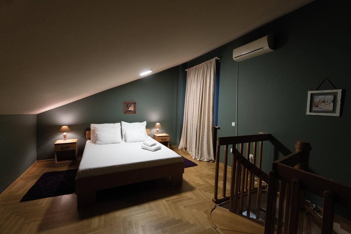 Ein Schlafzimmer mit einem Bett, zwei Nachttischen, einer Klimaanlage, einem Fenster mit Vorhängen und einem Holzbalken.