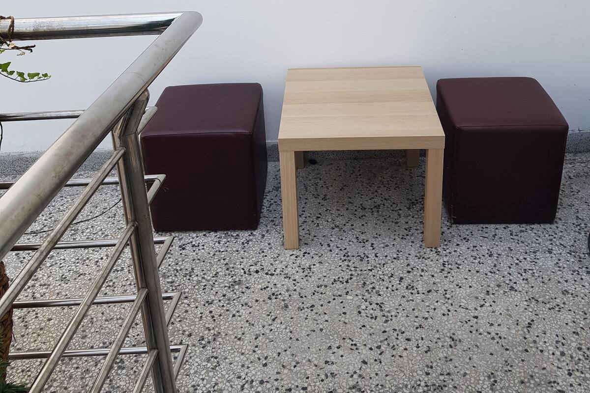Ein kleiner Sitzbereich im Freien mit einem Metallgeländer, einem Holztisch und zwei dunklen, gepolsterten Hocker.
