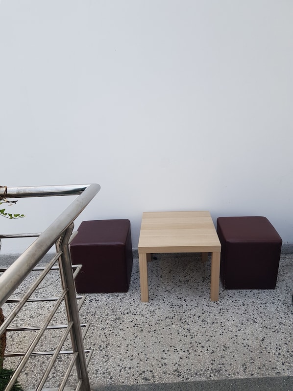 Uma pequena área externa com assentos, incluindo um corrimão metálico, uma mesa de madeira e dois banquinhos estofados e escuros.