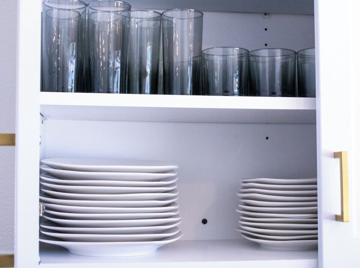 A white shelf with clear drinking glasses and white plates stacked on it.