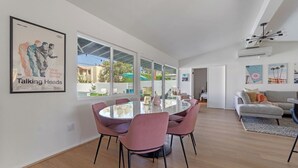 A modern living room with a round glass table and pink chairs, a grey sofa, and a large window with a view of the outside. - Poolside at Panorama - Walk to El Paseo (Palm Desert)