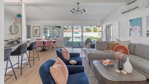 A modern living room with a sofa, chairs, and a coffee table. There is a dining area with chairs and a table. The room has large windows and a view of the outside. - Poolside at Panorama - Walk to El Paseo (Palm Desert)