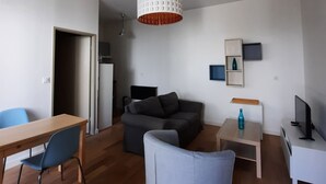 Ein modernes Wohnzimmer mit einem Holzesstisch, einem grauen Sofa, einem blauen Sessel, einer weißen Pendelleuchte und einem Fernseher auf einem weißen Lowboard.