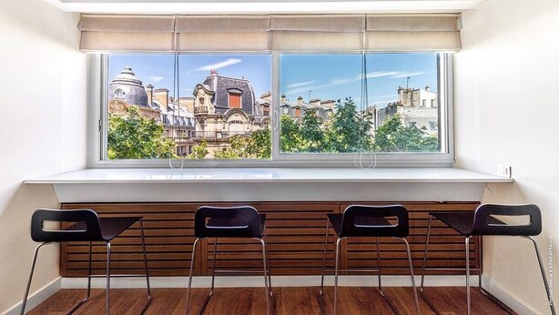 Una habitación con un amplio ventanal que da a un paisaje urbano, con listones de madera debajo de la ventana y tres sillas negras.