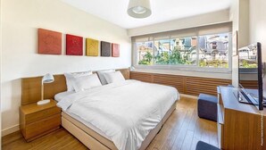 Un dormitorio moderno con una cama grande, cabecero de madera, mesitas de noche, un escritorio y un amplio ventanal con vistas a edificios.