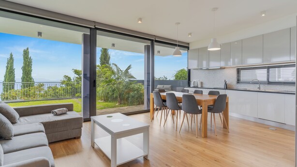 A modern kitchen with white cabinets, a dining area with a table and chairs, and a large sliding glass door leading to a balcony with a view of trees and a fence. - CR7 Airport View (Santa Cruz)