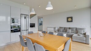 A modern kitchen with white cabinets, a built-in oven, and a dining area with a wooden table and grey chairs. - CR7 Airport View (Santa Cruz)