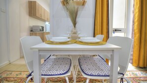 A dining table set with plates, glasses, and a vase with dried plants. There are two chairs with patterned cushions. A microwave and a shelf with plants are visible in the background. - Charming 2 bedroom townhouse, internal yard JDES1-1 (Gzira)