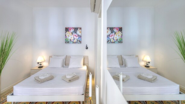 A hotel room with two beds, each with a bedside table and lamp, a floral painting on the wall, and a potted plant on the left side. - Charming 2 bedroom townhouse, internal yard JDES1-1 (Gzira)