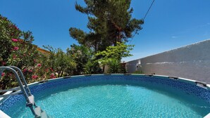 A clear blue swimming pool with a ladder, surrounded by lush greenery and a clear sky.